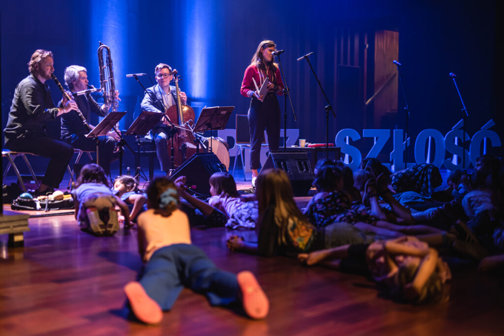 W tle grupa muzyków grająca na instrumentach. Z przodu grupka dzieci leżąca na scenie i słuchająca koncetru.