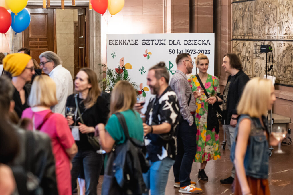 Grupa osób zgromadzona w holu poznańskiego Zamku. W tle baner z napisem: Biennale Sztuki dla Dziecka.