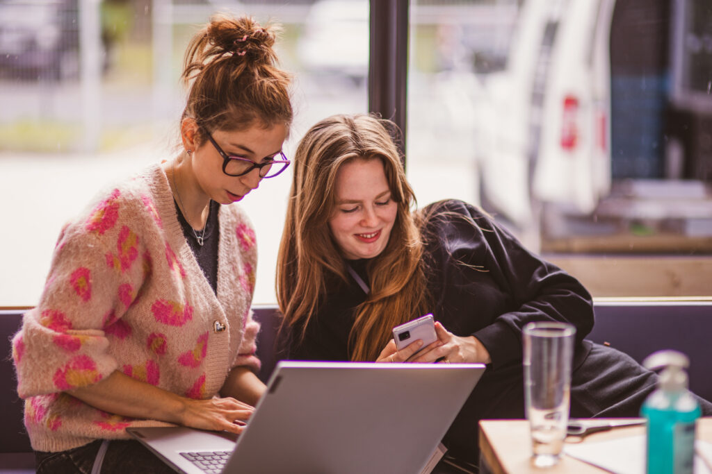 W budynku. Dwie kobiety siedzą przy stole, jedna z nich pracuje przy komputerze, druga z uśmiechem patrzy w telefon.