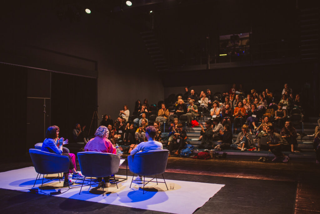 zdjęcie przedstawia cztery osoby siedzące na krzesłach na scenie, zwrócone przodem do publiczności i publiczność w tle