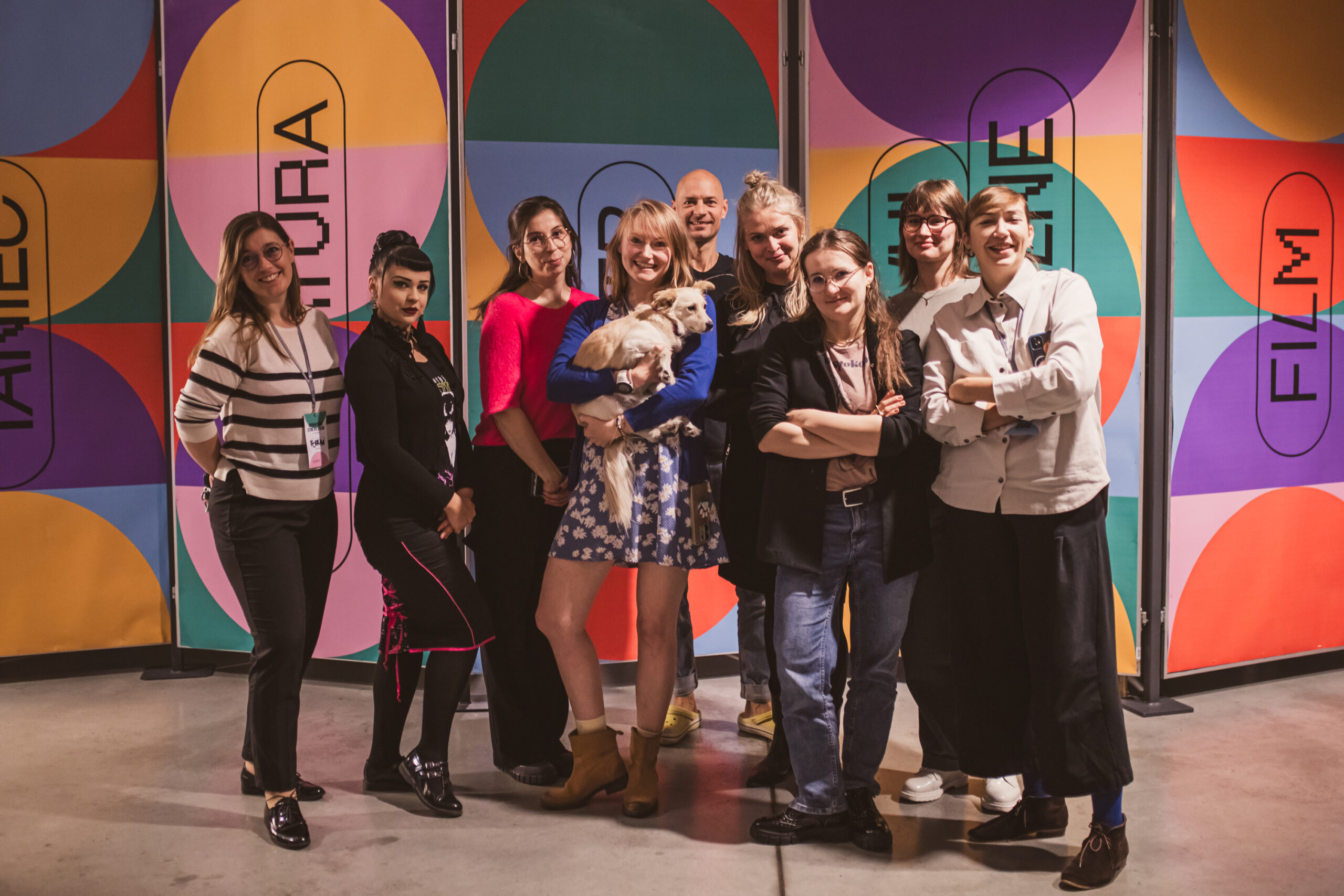 Grupa osób stoi ipozuje do zdjęcia. Uśmiechają się. Za nimi znajdują się kolorowe plakaty z napisami: teatr, sztuki wizualne. Jedna z osób, znajdująca się w centrum grupy trzyma na rękach niedużego pieska o jasnej sierści.