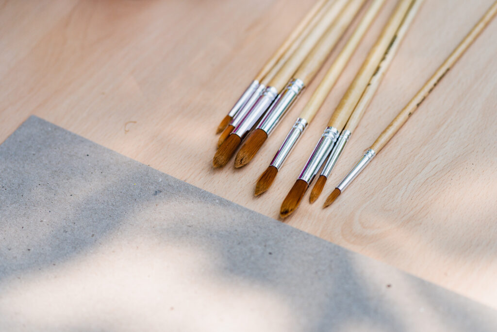 Zdjęcie przedstawia kartkę papieru i 8 leżących równolegle pędzli do malowania farbami.