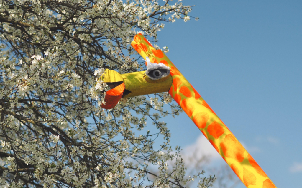 Zółto-pomarańczowa szyja żyrafy wykonana z rur z pianki na tle kwitnącego drzewa.