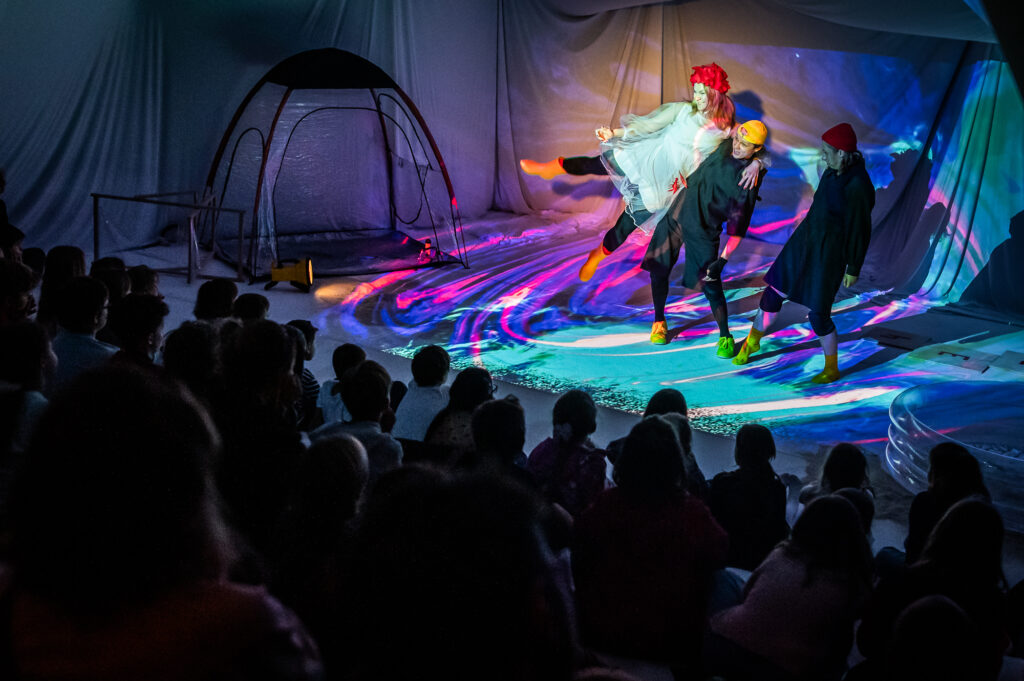 trójka aktorów na scenie z kolorowymi światłami, a na pierwszym planie publiczność