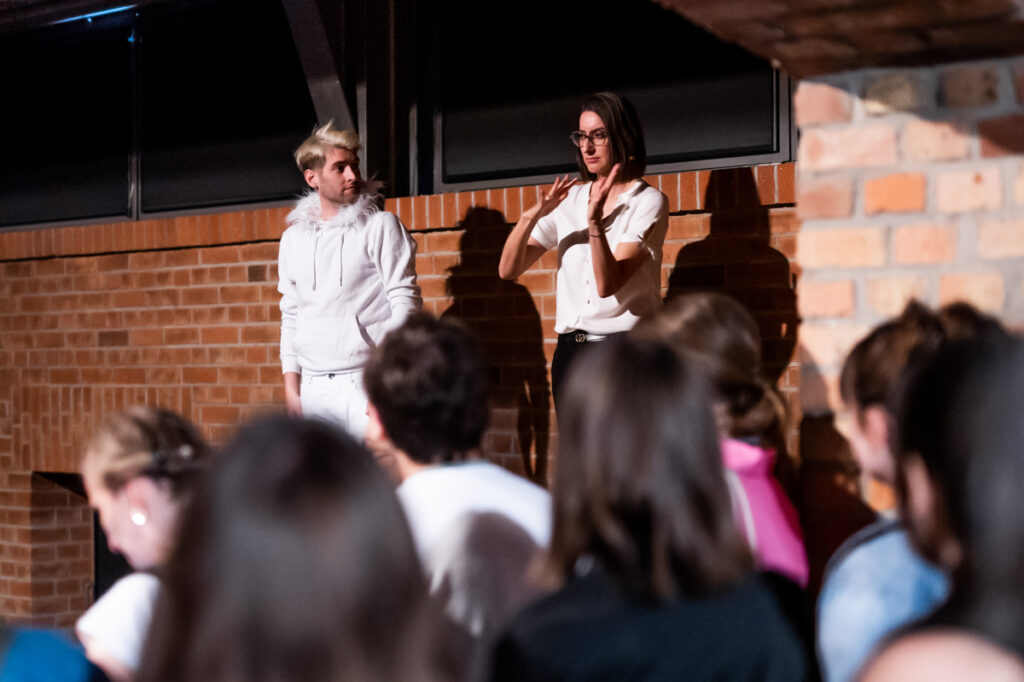 kobieta posługująca się językiem migowym na scenie, obok mężczyzna, publiczność w tle