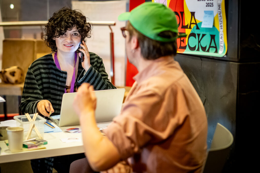 kobieta przed ekranem laptopa, rozmawia z mężczyzną odwróconym plecami